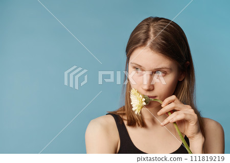 Medium close-up studio portrait of thoughtful young Caucasian woman with no makeup holding flower at her mouth looking away 111819219