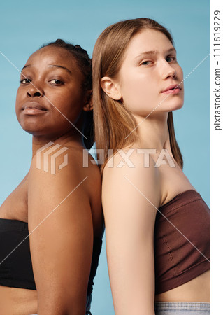 Vertical waist up studio portrait of two confident young Black and Caucasian women wearing bandeau tops standing back to back looking at camera 111819229