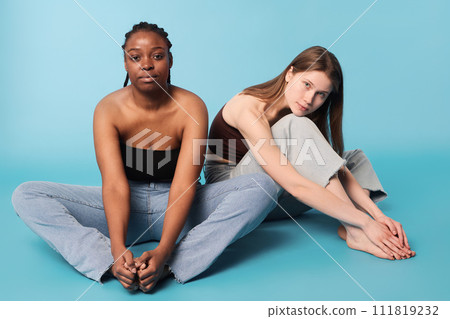 Two young ethnically diverse young women wearing casual clothes sitting relaxed on floor in studio looking at camera, sky blue background 111819232