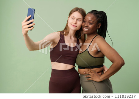 Medium studio portrait of two young ethnically diverse women wearing sportswear taking selfie on smartphone camera Medium studio portrait of two young ethnically diverse women wearing sportswear taking selfie on smartphone camera 111819237
