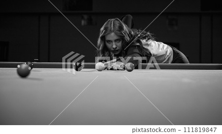 Focused young woman playing snooker game, aiming cue on balls. Online course banner for mastering billiards skills. Black and white Focused young woman playing snooker game, aiming cue on balls. Online course banner for mastering billiards skills. Black and white 111819847