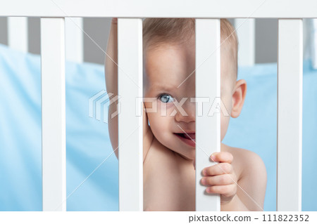 A baby in a playful mood looks slyly through the crib fence 111822352