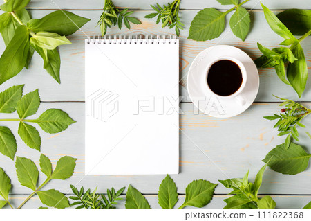 Spring composition with opened blank notepad, cup of black coffee, green leaves on wooden table. Morning coffee cup. 111822368