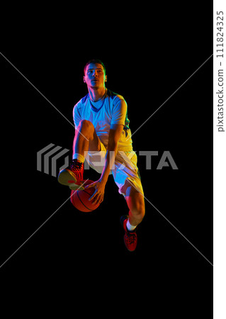 Young basketball athlete training dribbling technique in motion against black studio background in mixed neon light. Young basketball athlete training dribbling technique in motion against black studio background in mixed neon light. 111824325