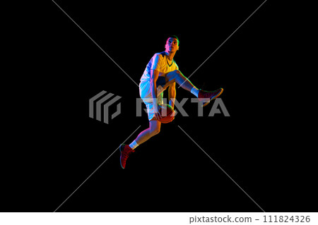 Dynamic portrait of young basketball player dribbling in mid-air against black studio background in mixed neon light. Dynamic portrait of young basketball player dribbling in mid-air against black studio background in mixed neon light. 111824326
