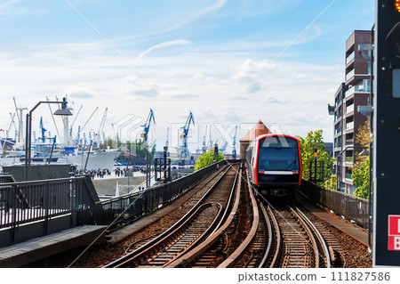 Hamburg Red U-bahn Subway train departure Baumwall station on Elevated Track with Hamburger Port Cranes and ships cargo harbor background blue sky. Hanseatic city commute transport scenic view 111827586
