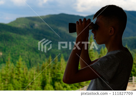 Boy teenager looks through binoculars at the mountains. 111827709