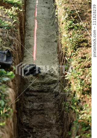 An orange reinforced sewer pipe is compacted with sand in a trench. Plumbing works on private plot. An orange reinforced sewer pipe is compacted with sand in a trench. Plumbing works on private plot. 111828239