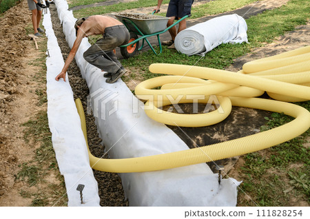 Man pours rubble from a wheelbarrow into a trench. Drainage works for drainage of ground water around the field. Man pours rubble from a wheelbarrow into a trench. Drainage works for drainage of ground water around the field. 111828254