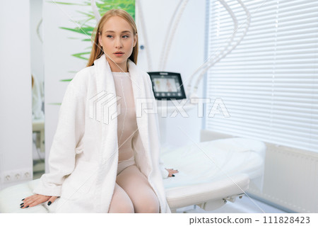 Portrait of pretty young blonde female patient in white bath robe sitting on medical couch in modern cosmetology clinic while waiting beautician specialist. Concept of cosmetology, beauty treatment. Portrait of pretty young blonde female patient in white bath robe sitting on medical couch in modern cosmetology clinic while waiting beautician specialist. Concept of cosmetology, beauty treatment. 111828423