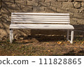 A white wooden bench stands against an old brick wall, on the ground with dried autumn grass and leaves, illuminated by the bright sun 111828865