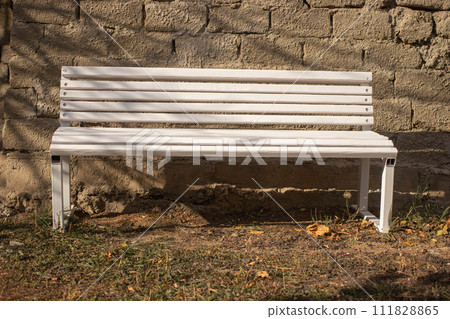 A white wooden bench stands against an old brick wall, on the ground with dried autumn grass and leaves, illuminated by the bright sun A white wooden bench stands against an old brick wall, on the ground with dried autumn grass and leaves, illuminated by the bright sun 111828865