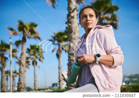Determined young female athlete, sportswoman holding a steel bottle with refreshing water, relaxing after run outdoor Determined young female athlete, sportswoman holding a steel bottle with refreshing water, relaxing after run outdoor 111830887