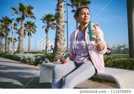 Attractive sporty woman in activewear, sitting on a bench on the marine promenade, drinking water after workout or run 111830889
