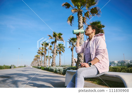 Active female athlete drinks water from a bottle, resting after intense workout, sitting on a stone bench in public park 111830908