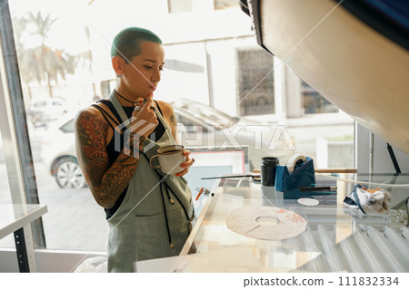 Focused female worker drink coffee while working with printing machine in a workshop 111832334
