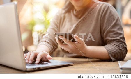 A woman multitasking, using her smartphone and laptop computer, working remotely at a cafe. A woman multitasking, using her smartphone and laptop computer, working remotely at a cafe. 111835828