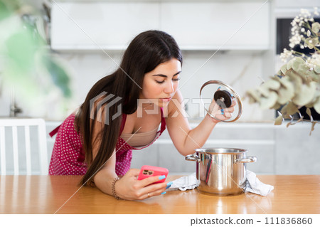 Young woman in apron with phone looks at pan 111836860