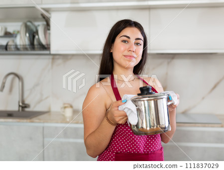 Smiling young woman wearing apron holding hot pan in the kitchen 111837029