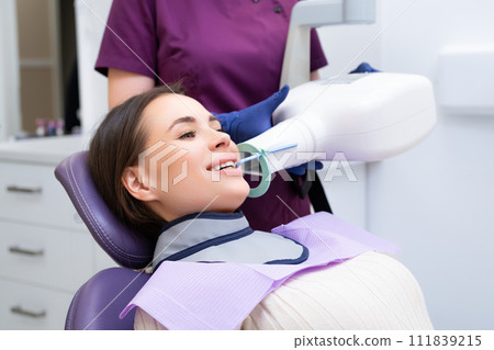 Dental assistant prepares patient for X-ray using specialized tools in clinic. Woman asks female patient to smile for better view of mouth Dental assistant prepares patient for X-ray using specialized tools in clinic. Woman asks female patient to smile for better view of mouth 111839215