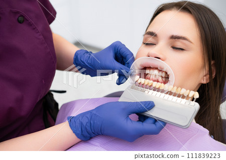 The dentist carefully selects the enamel shade for the young brunette womans teeth before beginning the whitening procedure. The dentist carefully selects the enamel shade for the young brunette womans teeth before beginning the whitening procedure. 111839223