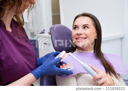 Dental professional explains to female patient how to brush teeth correctly in clinic. Dentists uses human jaw dental model for clarity Dental professional explains to female patient how to brush teeth correctly in clinic. Dentists uses human jaw dental model for clarity 111839225