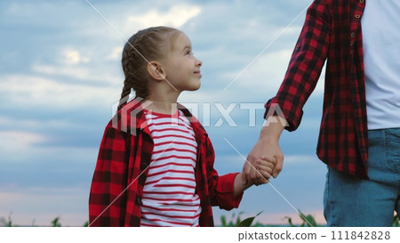 Girl ambles through corn field hand in hand with farmer mom. Daughter has weekend with mother guided by hand through corn field. Girl and agriculturist spend time together among corn field Girl ambles through corn field hand in hand with farmer mom. Daughter has weekend with mother guided by hand through corn field. Girl and agriculturist spend time together among corn field 111842828