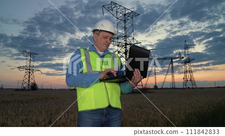 Man worker conducts examination of power transmission lines with laptop in sunset rural field. Mature engineer with laptop works at power generation substation at evening countryside. Power system Man worker conducts examination of power transmission lines with laptop in sunset rural field. Mature engineer with laptop works at power generation substation at evening countryside. Power system 111842833