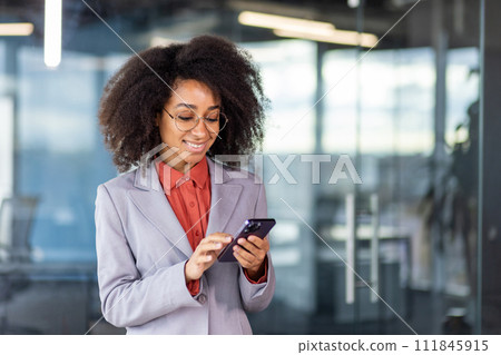 Satisfied executive in corporate dress code using modern smartphone while standing in cabinet with glassy transparent walls. Careless woman scrolling news in wireless gadget during break from work. 111845915