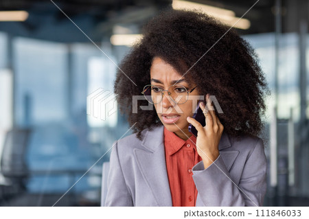 Worried african american lady with bushy hairstyle holding smartphone by ear and listening bad news. Disturbed office worker having stressful distant conversation by mobile gadget during daytime. 111846033