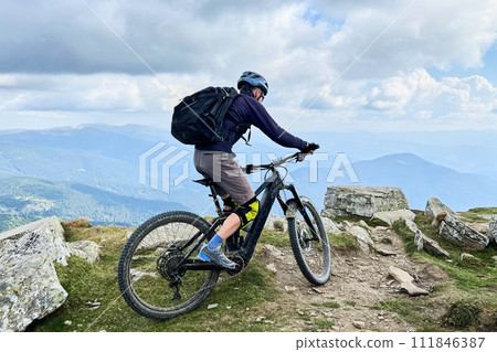 Cyclist man riding electric mountain bike outdoors. Male tourist biking along grassy trail in the mountains, wearing helmet and backpack. Concept of sport, active leisure and nature. Cyclist man riding electric mountain bike outdoors. Male tourist biking along grassy trail in the mountains, wearing helmet and backpack. Concept of sport, active leisure and nature. 111846387