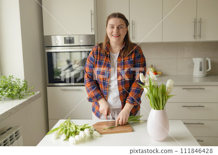 Happy Caucasian 30s woman taking care of cut fresh bouquet of tulips in kitchen. Housewife hands of woman cutting flower on wooden board Happy Caucasian 30s woman taking care of cut fresh bouquet of tulips in kitchen. Housewife hands of woman cutting flower on wooden board 111846428