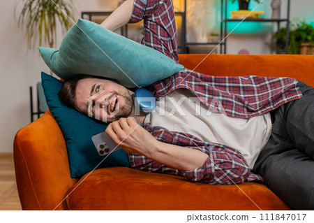 Irritated angry man covers ears with cushions annoyed by noisy loud music neighbors repair work Irritated angry man covers ears with cushions annoyed by noisy loud music neighbors repair work 111847017