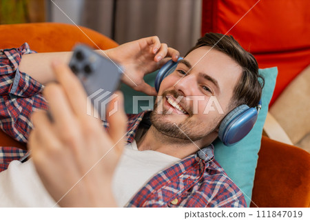 Man in wireless headphones listening energetic dancing music on smartphone relaxing lying on sofa 111847019