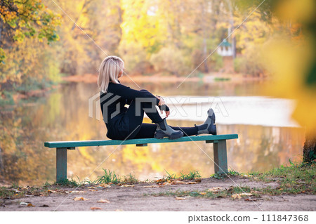 An attractive blonde woman with an athletic build rests sitting on a bench in a park near a lake and looks at a beautiful sunny autumn sunset An attractive blonde woman with an athletic build rests sitting on a bench in a park near a lake and looks at a beautiful sunny autumn sunset 111847368