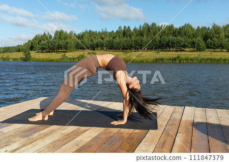 Attractive woman performing Setu Bandhasana exercise, bridge pose, working out in leggings and a short top on the shore of a lake on a sunny warm morning Attractive woman performing Setu Bandhasana exercise, bridge pose, working out in leggings and a short top on the shore of a lake on a sunny warm morning 111847379