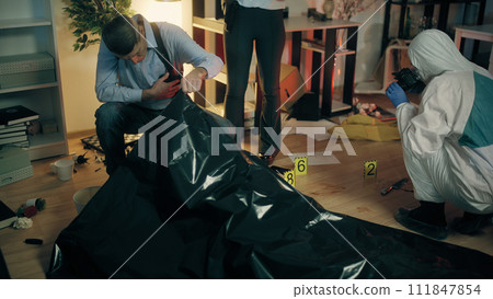 A male detective is examining a dead body while his colleague looks at the evidence A male detective is examining a dead body while his colleague looks at the evidence 111847854
