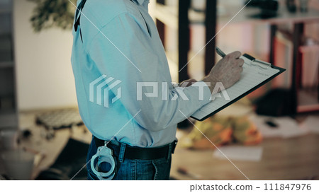 A police detective is filling out report papers while examining the crime scene in an apartment A police detective is filling out report papers while examining the crime scene in an apartment 111847976