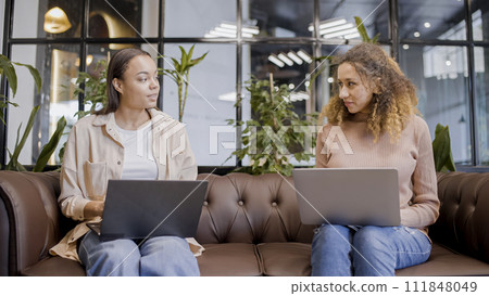 Two women talking, typing on laptops in office, female IT specialists at work Two women talking, typing on laptops in office, female IT specialists at work 111848049