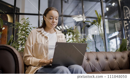 Smiling game developer working on laptop, sitting on sofa in office, education 111848050