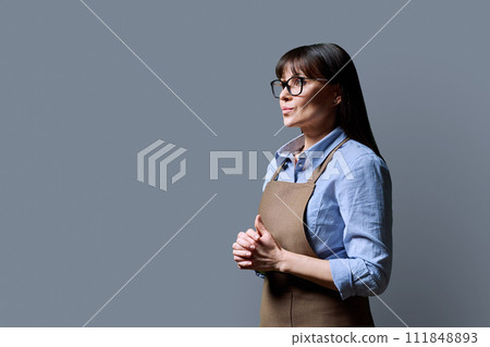 Middle-aged confident woman in apron looking in profile on gray background Middle-aged confident woman in apron looking in profile on gray background 111848893