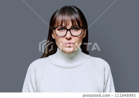 Headshot portrait middle-aged serious woman in white sweater on gray background 111848993