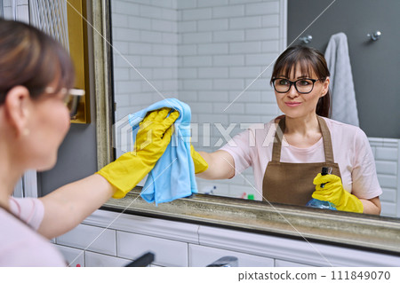 Middle-aged woman in an apron cleaning mirror in bathroom Middle-aged woman in an apron cleaning mirror in bathroom 111849070