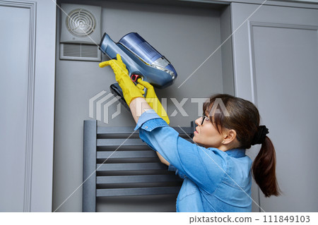 Woman cleaning wall-mounted hood ventilation grill in bathroom with vacuum cleaner 111849103