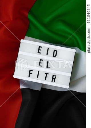 EID EL FITR - Eid Mubarak - Happy Holidays text frame on United Arab Emirates waving flag made from silk material. Public holiday celebration background. The National Flag of UAE. Commemoration Day 111849345