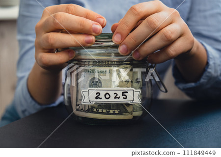 Female hands holding Glass jar full of American currency dollars cash banknote with text 2025 year. Preparation saving money. Moderate consumption and economy Female hands holding Glass jar full of American currency dollars cash banknote with text 2025 year. Preparation saving money. Moderate consumption and economy 111849449
