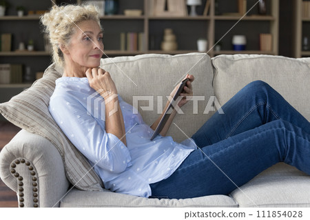 Pensive older woman using tablet device seated on comfortable sofa 111854028