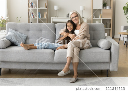 Positive pretty caring grandmother hugging girl on soft trendy couch Positive pretty caring grandmother hugging girl on soft trendy couch 111854513