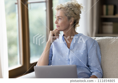Serious older woman looking away sit on sofa with laptop 111854805