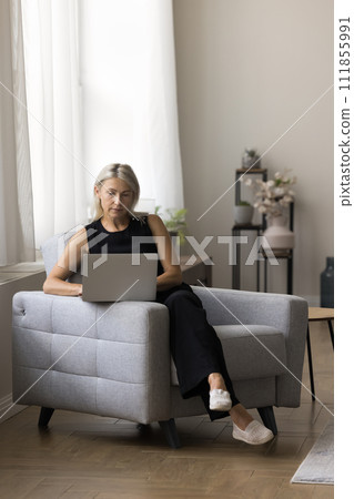 Serious mature woman checks email sitting on armchair with laptop 111855991